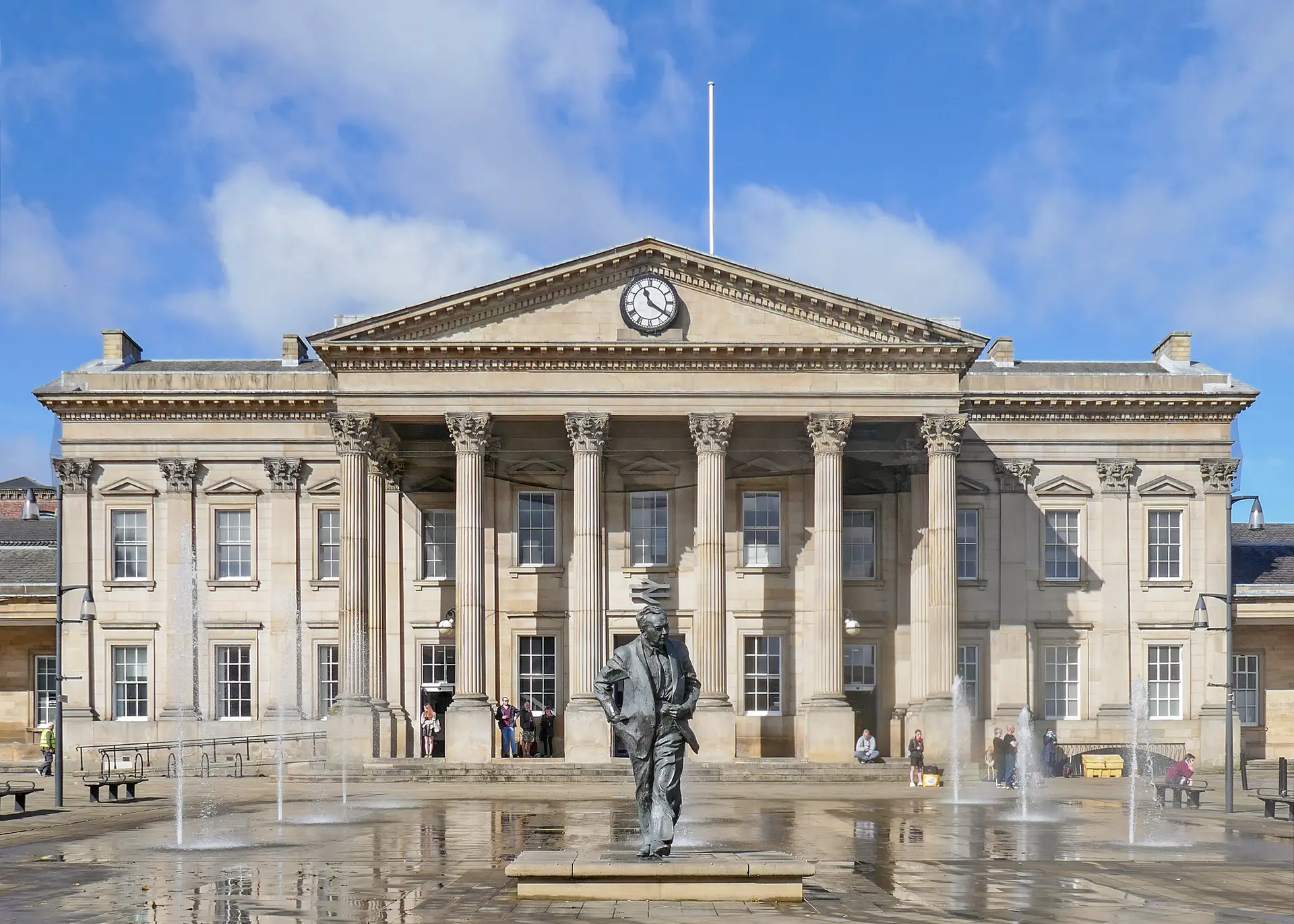 The classical colonnaded front of Huddersfield railway station on St George's Square, with fountains and the Harold Wilson statue