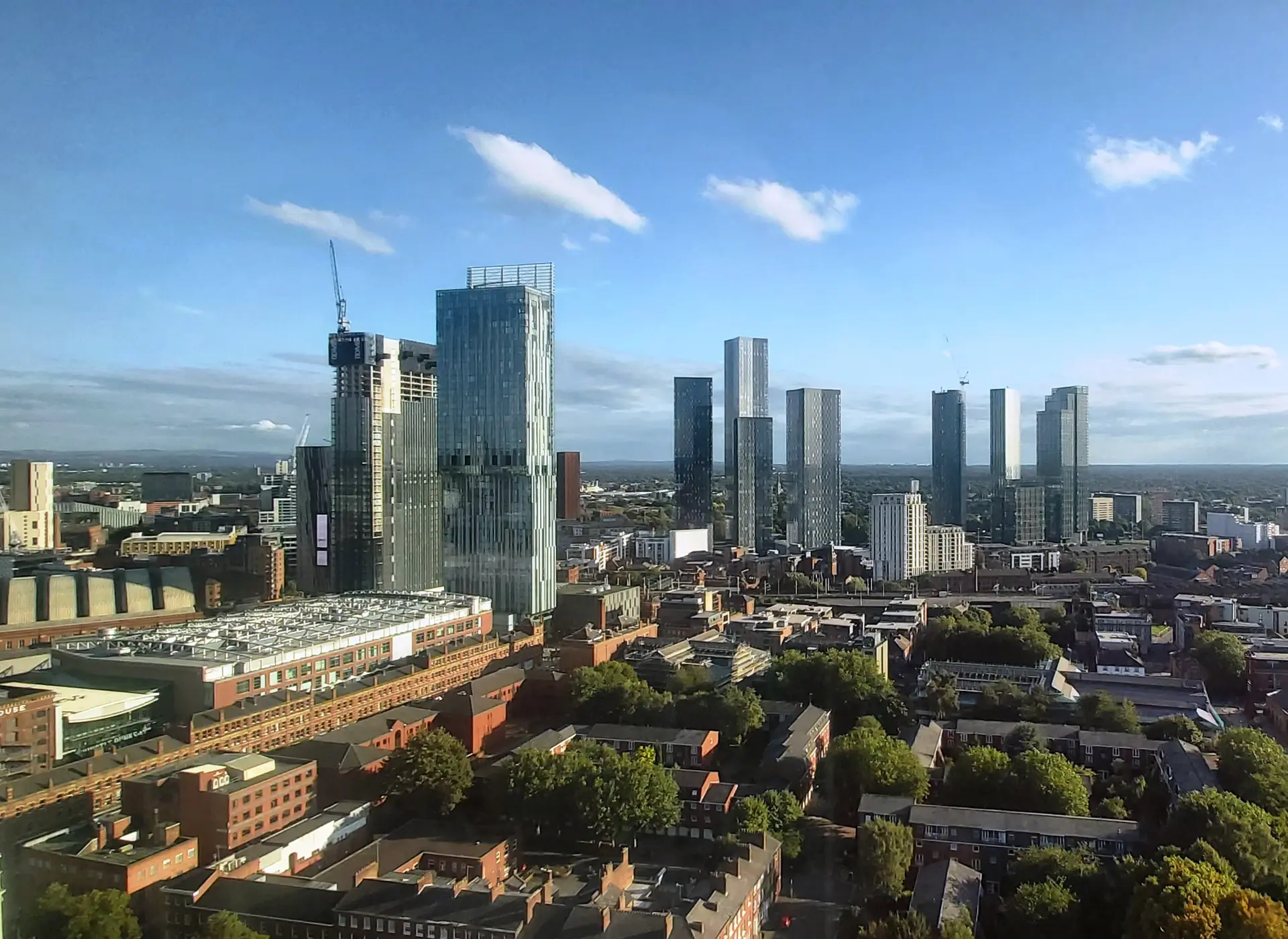 Manchester city-centre skyline, with glass towers rising above red-brick buildings and trees