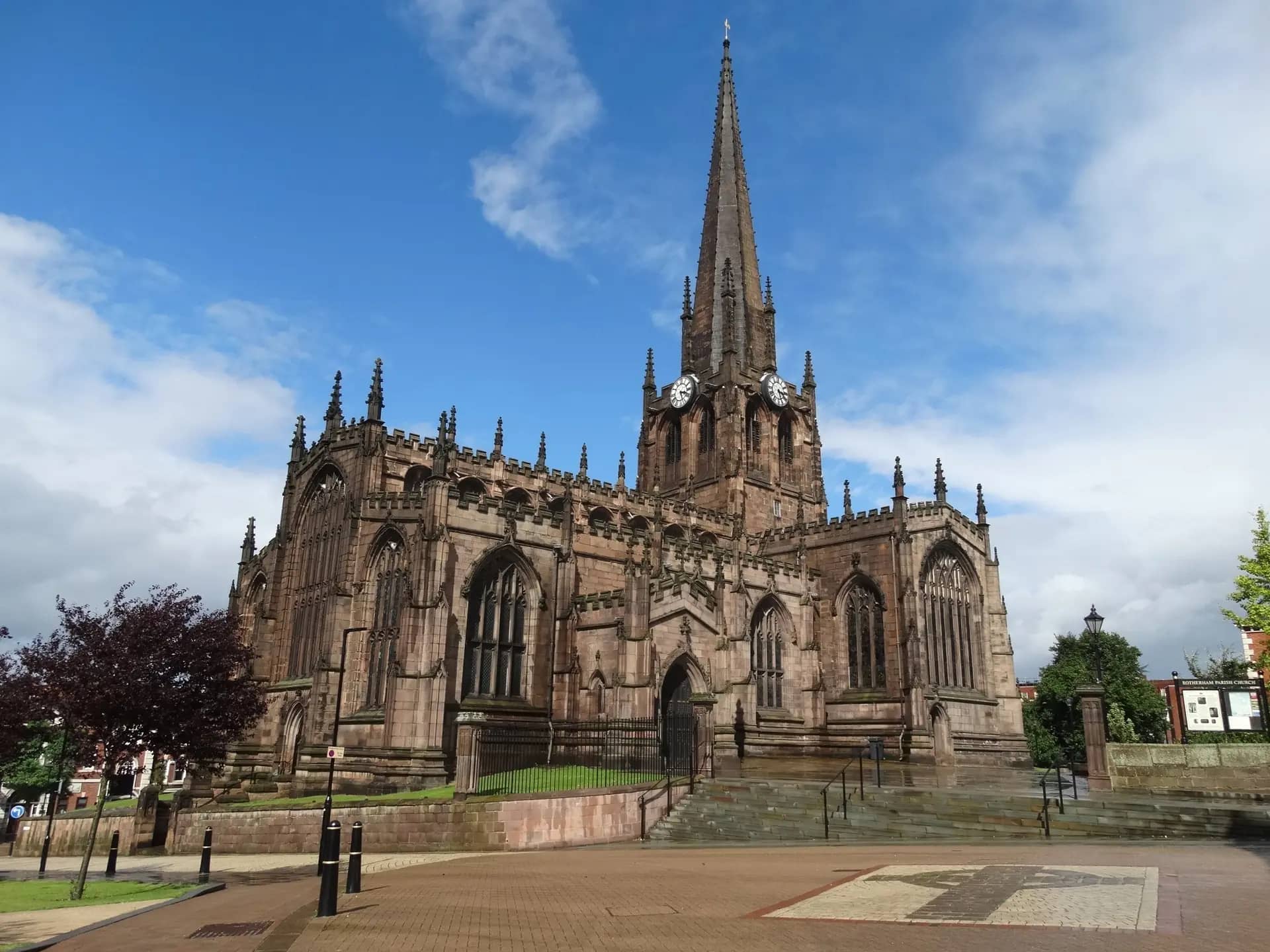 The spire and sandstone nave of Rotherham Minster under a blue sky, seen across the paved square