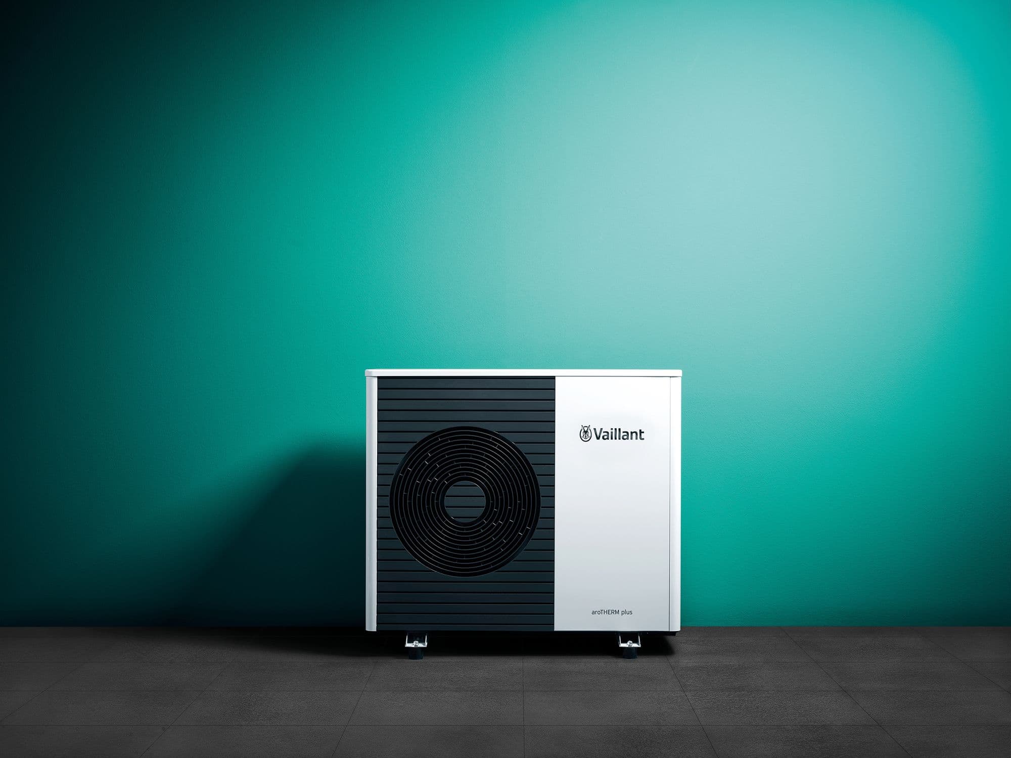 Vaillant aroTHERM plus outdoor unit stood on a tiled floor against a plain green wall, fan grille facing the camera