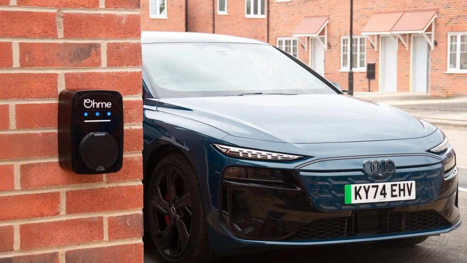 An Ohme home charger on a brick wall beside a parked electric car