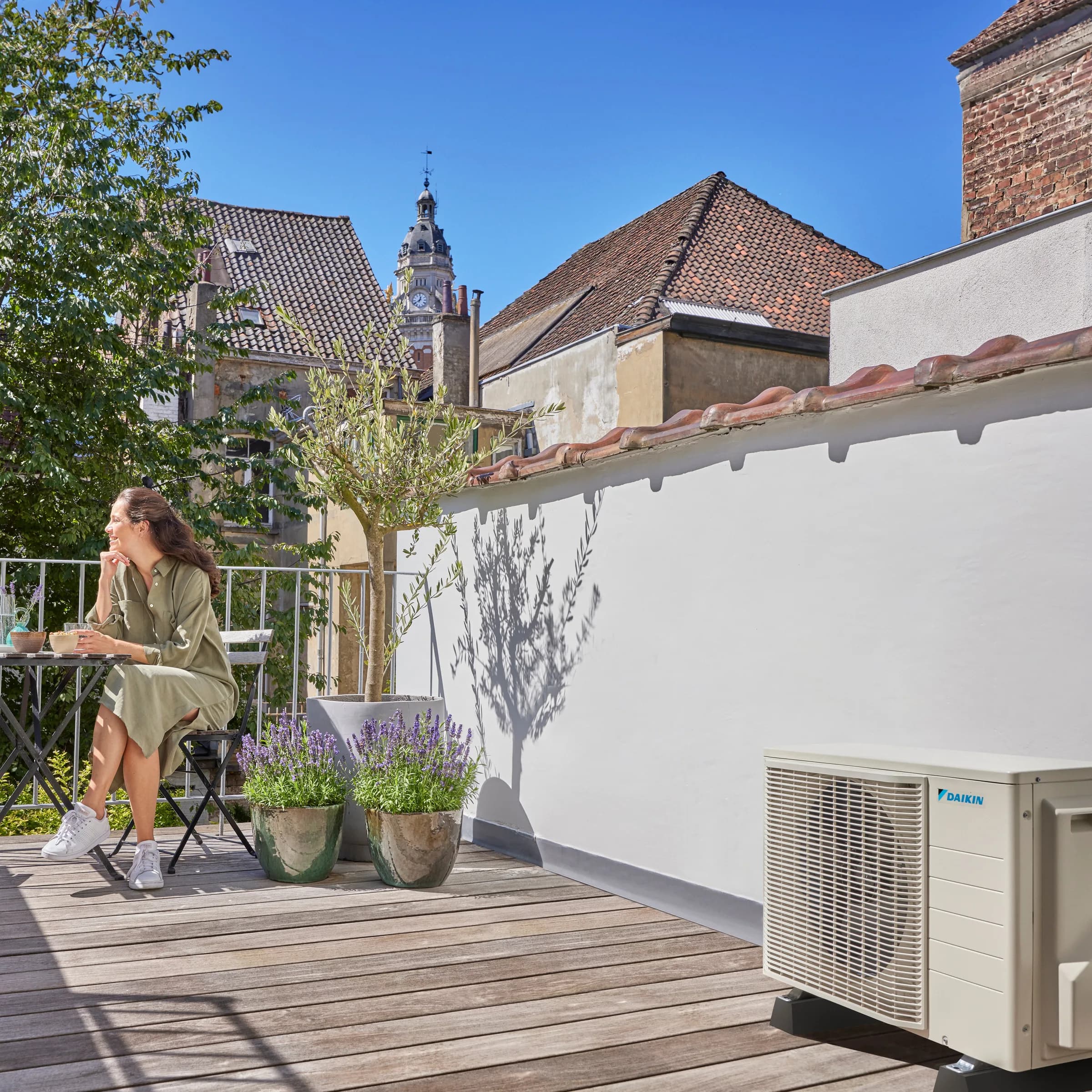 A Daikin outdoor unit on a terrace beside a house, with a woman sitting out at a table
