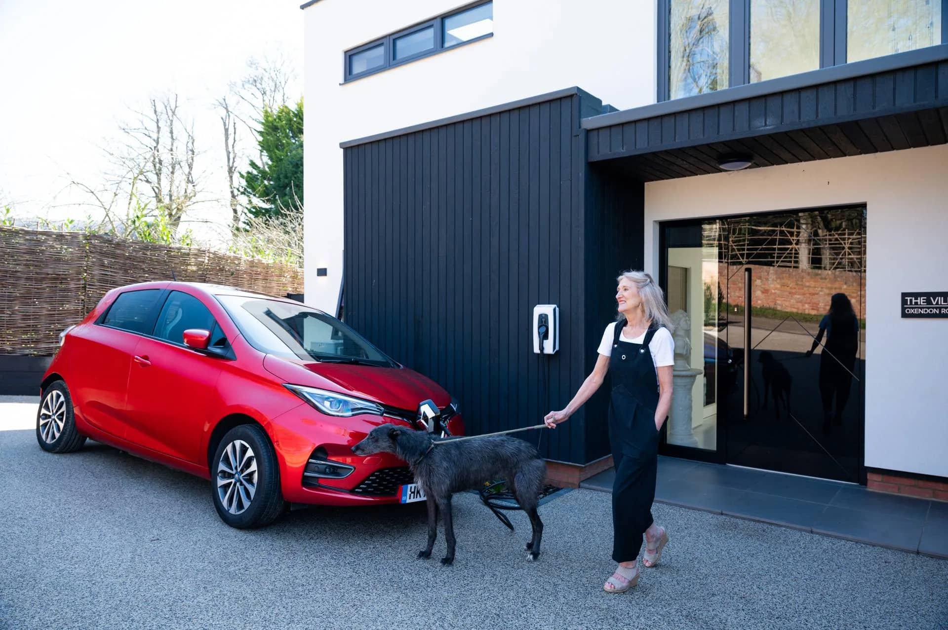 A woman walking a dog past an electric car charging outside a house