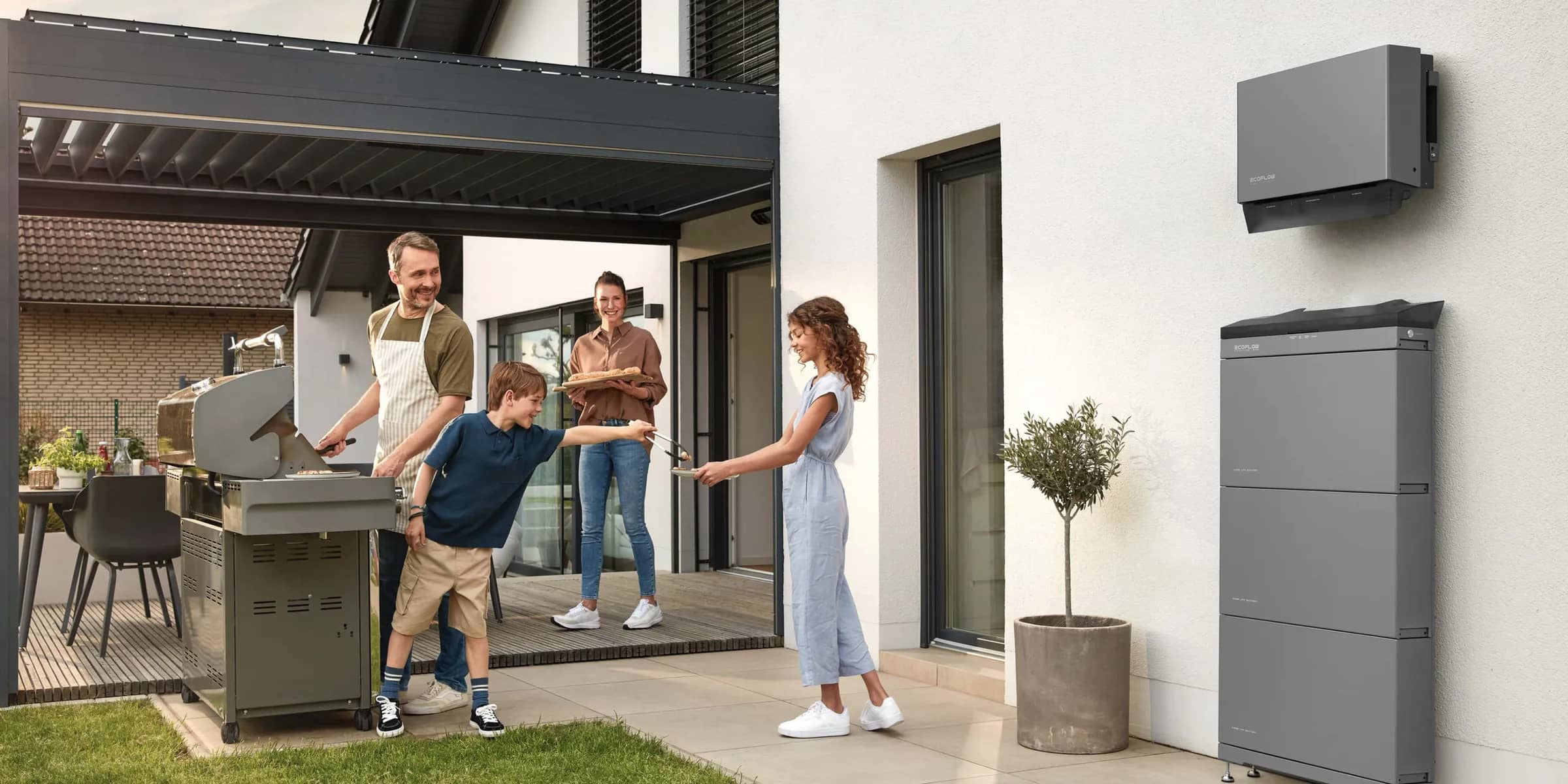 A family at a barbecue on the patio, with a home battery and inverter on the wall behind them