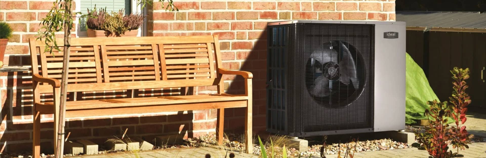 An Ideal Logic Air heat pump against a brick wall beside a wooden garden bench