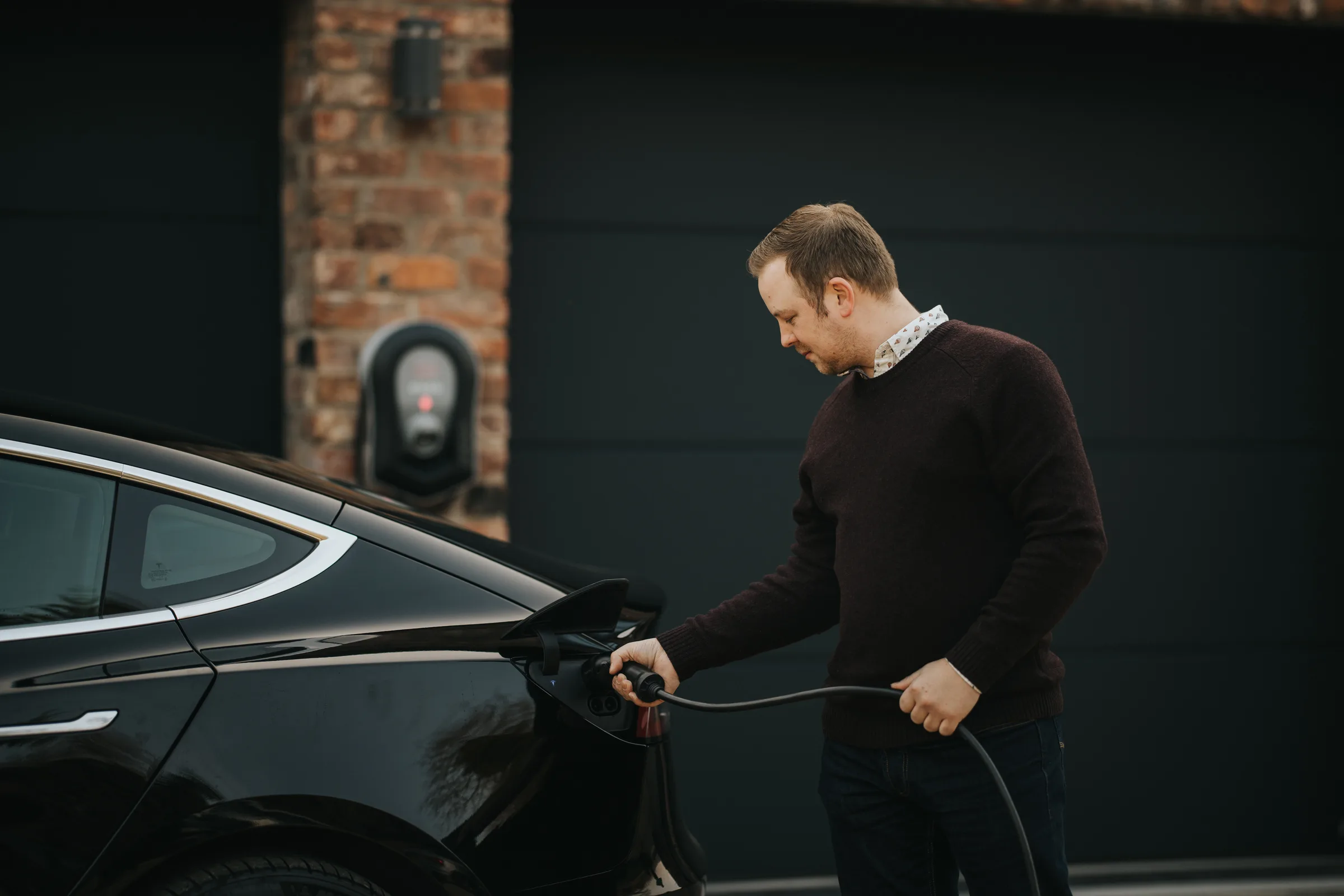A man plugging a charging cable into an electric car on a drive after dark
