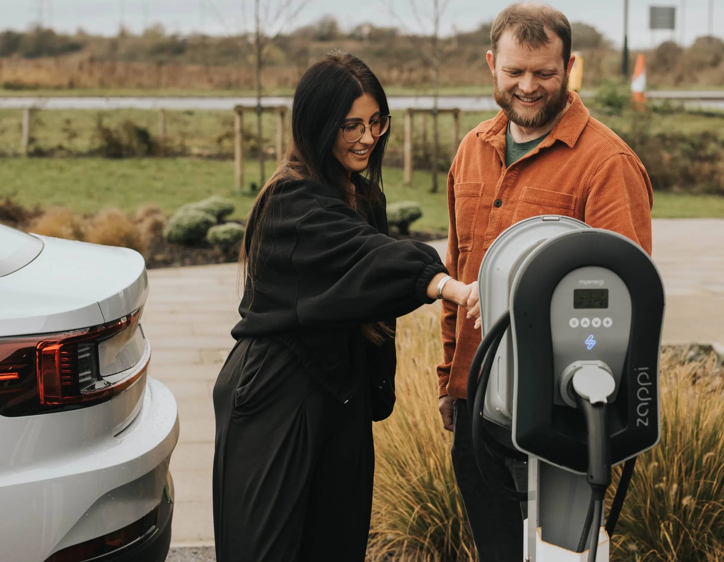 A couple laughing beside a myenergi Zappi charger on a post, with their car parked next to it